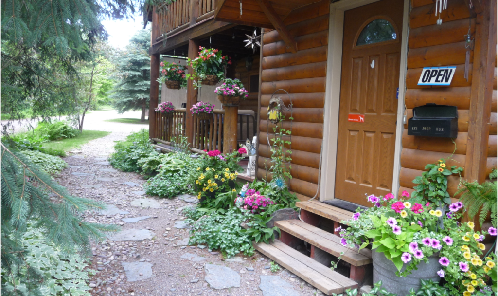 gravel pathway with log cabin. Flowers are in bloom and there is a open sign on the cabin door.